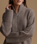 A close-up of a woman wearing a taupe brown ribbed knit half-zip sweater with a high mock neck collar, set against a solid warm beige background.