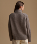 A woman shown from the back wearing a taupe brown ribbed knit turtleneck sweater and cream pants, set against a solid warm beige studio background.