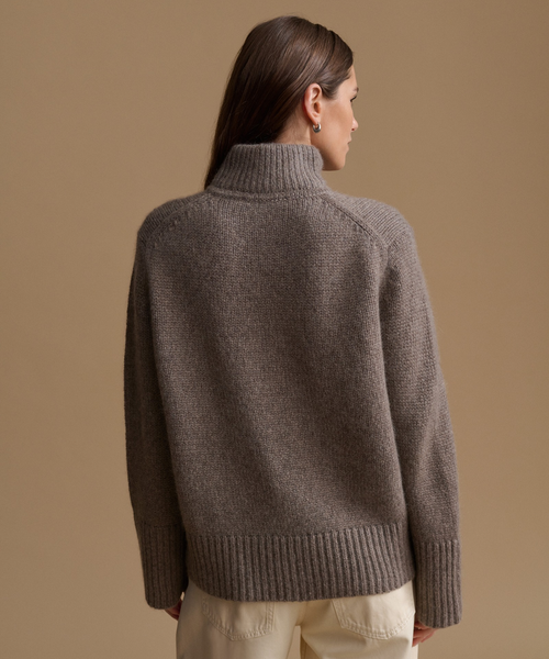 A woman shown from the back wearing a taupe brown ribbed knit turtleneck sweater and cream pants, set against a solid warm beige studio background.