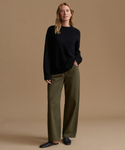 A woman stands against a plain warm beige studio background wearing a black knit sweater, olive green wide-leg trousers, and dark flat shoes.