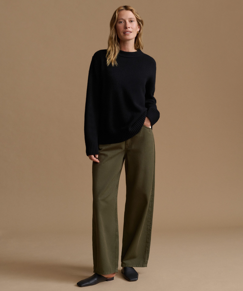A woman stands against a plain warm beige studio background wearing a black knit sweater, olive green wide-leg trousers, and dark flat shoes.