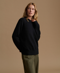 A woman modeling a black ribbed knit sweater with a mock neck, paired with olive green trousers, set against a solid warm beige studio background.