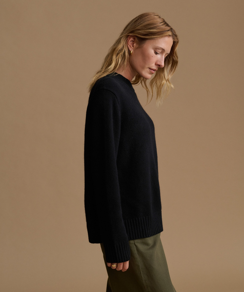 A woman wearing a black knit sweater and olive green pants, shown in profile against a solid warm beige studio background.