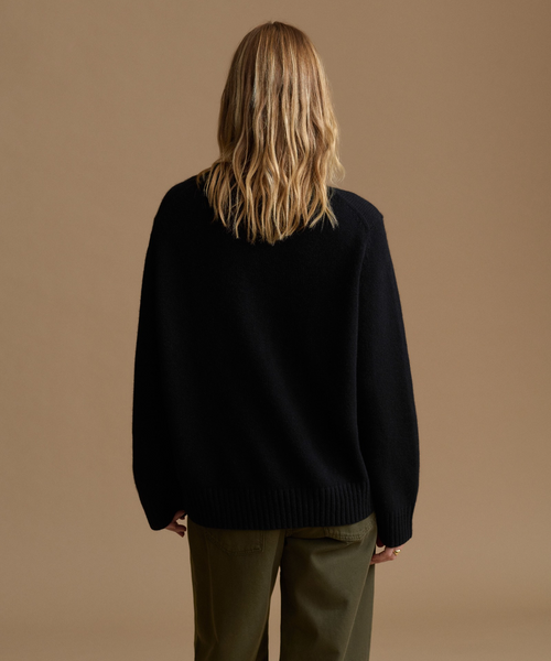 A woman viewed from behind wearing a black knit sweater and olive green trousers, standing against a solid warm beige studio background.