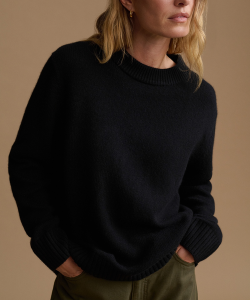 A woman wearing a black knit crewneck sweater with ribbed cuffs and collar, paired with olive green pants, against a solid warm beige background.