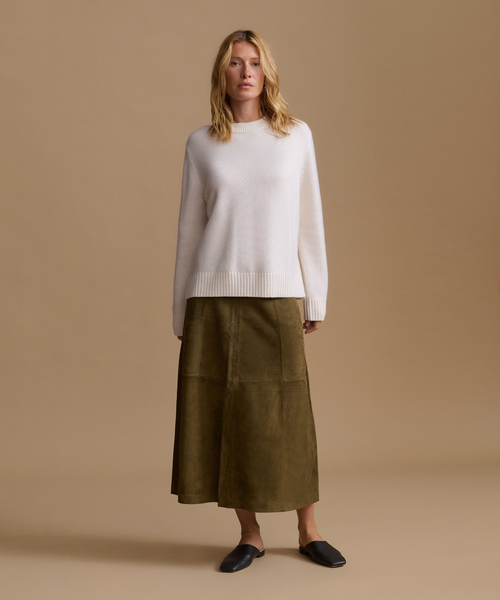 A woman stands against a plain warm beige studio background wearing a cream knit sweater, an olive green midi skirt, and black flat shoes.