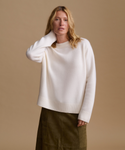 A woman modeling a cream-colored knit sweater with ribbed trim, paired with an olive green skirt, against a solid beige background.