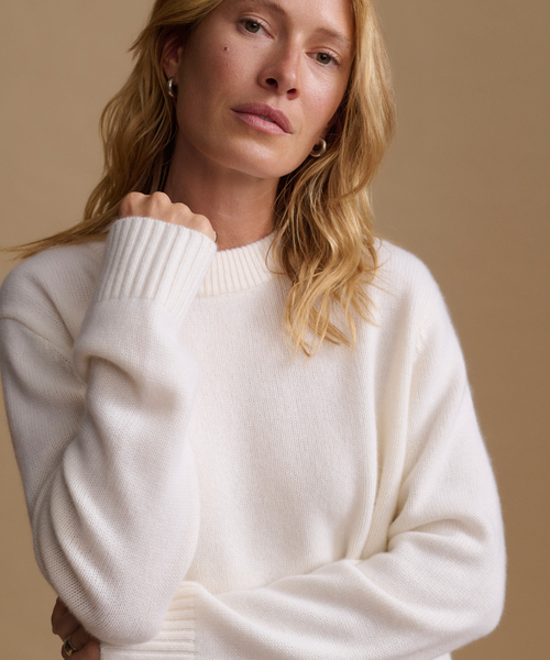 A close-up of a woman wearing a cream-colored, ribbed-knit crewneck sweater against a solid beige background.