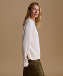 A woman wearing a cream knit sweater and an olive green skirt, photographed from the side against a solid beige studio background.