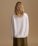 A woman viewed from behind wearing a cream-colored knit sweater and olive green trousers, set against a solid warm beige background.