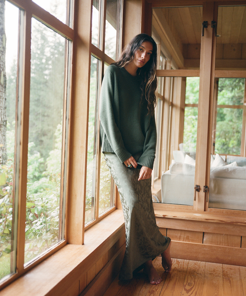 A woman stands barefoot on a wooden floor in a sunroom, wearing an oversized dark green knit sweater and a matching textured green maxi skirt. Large windows with wood frames look out onto a wooded outdoor scene, and a mirror reflects a light-colored sofa in the room.
