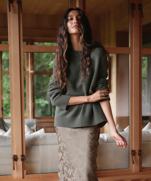 A woman stands indoors in front of large wooden-framed windows, wearing an oversized dark olive green knit sweater and an olive patterned skirt.