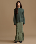 A woman stands against a plain warm beige studio background wearing a dark green knit sweater and a matching green maxi skirt, with dark flats.