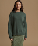 A woman modeling an oversized crewneck knit sweater in a muted sage green, paired with a matching textured skirt, against a solid tan studio background.
