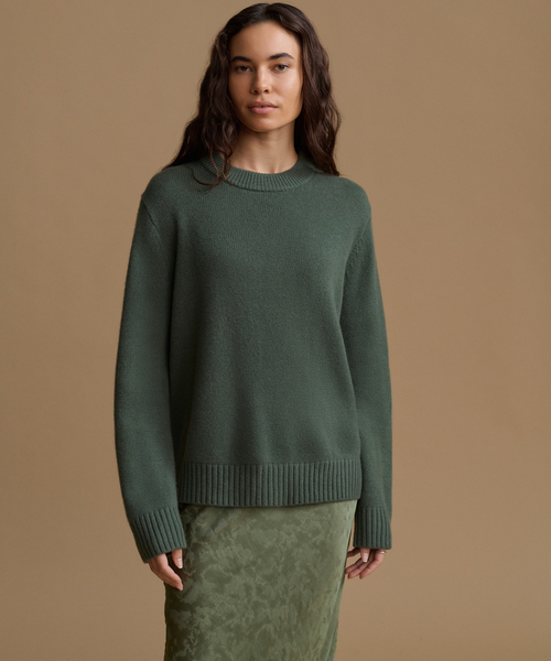 A woman modeling an oversized crewneck knit sweater in a muted sage green, paired with a matching textured skirt, against a solid tan studio background.