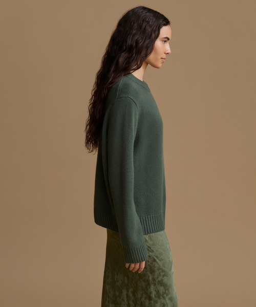 A woman in side profile wearing a dark green knit sweater and a matching green skirt, photographed against a plain warm beige studio background.