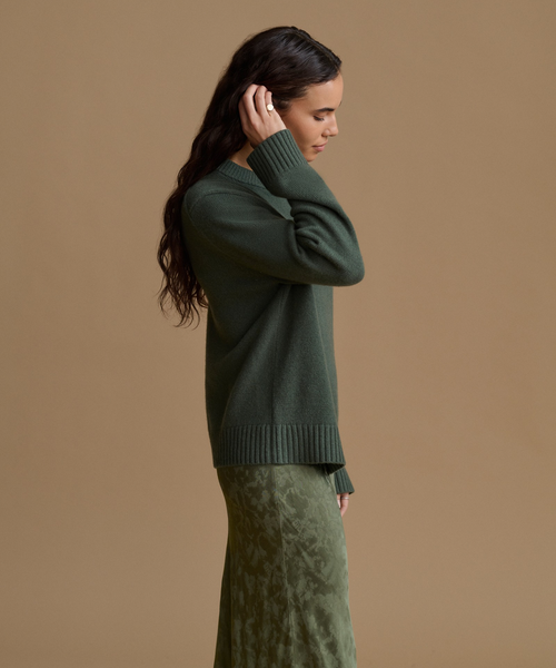 A woman in profile wearing a dark green knit sweater and a matching green skirt, standing against a solid tan background.