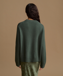 A woman shown from the back wearing a dark forest green knit sweater and a textured olive green skirt, set against a solid warm tan studio background.