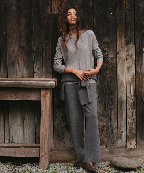 A woman stands against a rustic dark wood plank wall wearing a light gray knit long-sleeve top and matching gray wide-leg trousers, with brown shoes. A wooden bench is visible to her left.