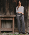 A woman stands outdoors against a dark wooden plank wall, wearing a light gray long-sleeve knit top, matching gray wide-leg pants, and a gray wrap tied around her waist. A rustic wooden table is to her left, and gravel is visible at the bottom.
