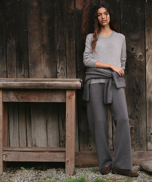 A woman stands outdoors against a dark wooden plank wall, wearing a light gray long-sleeve knit top, matching gray wide-leg pants, and a gray wrap tied around her waist. A rustic wooden table is to her left, and gravel is visible at the bottom.
