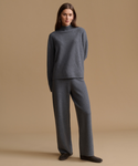 A woman modeling a matching dark gray knit lounge set consisting of a mock neck sweater and wide-leg trousers, standing against a solid beige studio background.