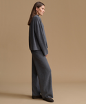 A woman stands in profile wearing a matching dark gray knit lounge set consisting of a cropped long-sleeve top and wide-leg pants, paired with dark slip-on shoes, against a solid beige studio background.