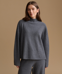 A woman wearing a dark gray knit mock neck sweater and matching knit pants, standing against a solid beige background.