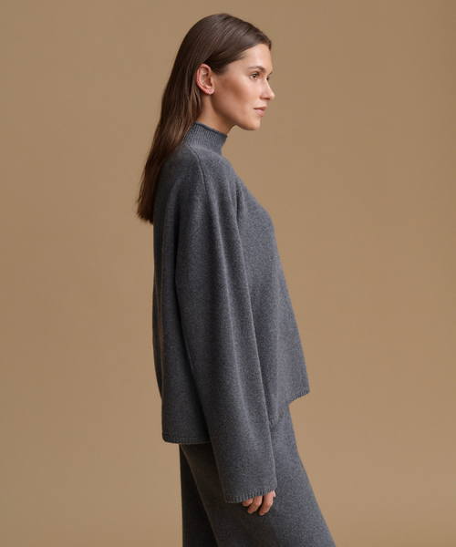 A woman in side profile wearing a dark gray knit mock-neck sweater and matching knit skirt against a solid tan background.