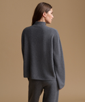 A woman shown from the back wearing a dark gray knit mock-neck sweater and matching gray trousers, standing against a solid warm beige studio background.