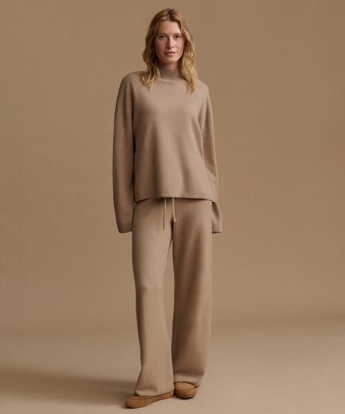 A woman stands against a warm beige studio background wearing a matching knit lounge set consisting of a long-sleeve mock-neck sweater and wide-leg trousers in a light camel color, paired with tan flat shoes.