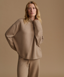 A woman modeling an oversized knit sweater and matching knit pants in a warm taupe-beige color, set against a solid matching beige studio background.