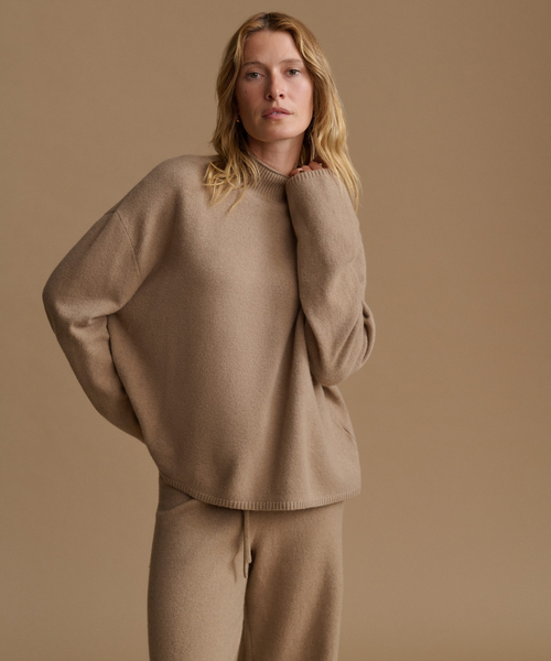A woman modeling an oversized knit sweater and matching knit pants in a warm taupe-beige color, set against a solid matching beige studio background.