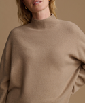 A close-up of a woman wearing an oversized ribbed mock neck sweater in a warm camel beige color, set against a matching solid beige background.