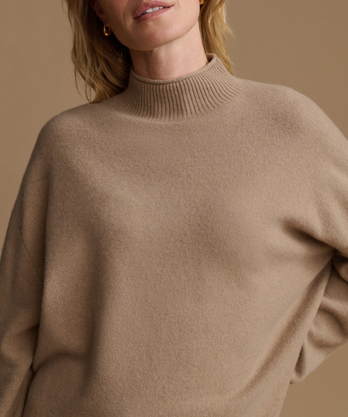 A close-up of a woman wearing an oversized ribbed mock neck sweater in a warm camel beige color, set against a matching solid beige background.