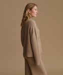 A woman in side profile wearing a matching beige knit sweater and trousers against a solid beige studio background.