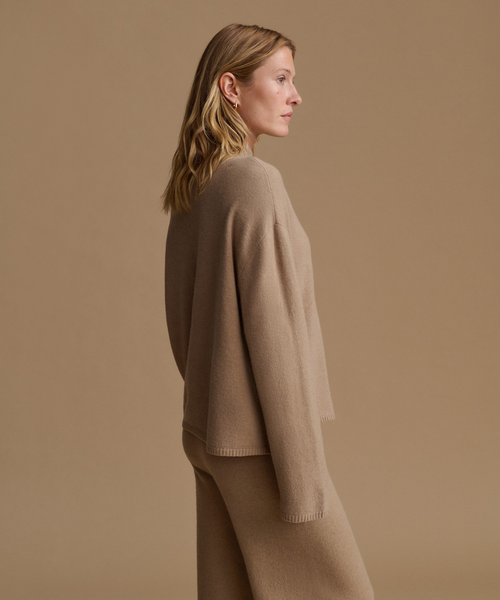 A woman in side profile wearing a matching beige knit sweater and trousers against a solid beige studio background.