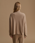 A woman shown from the back wearing a loose, light brown knit mock-neck sweater and matching knit pants against a solid warm beige background.