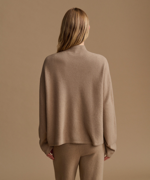 A woman shown from the back wearing a loose, light brown knit mock-neck sweater and matching knit pants against a solid warm beige background.