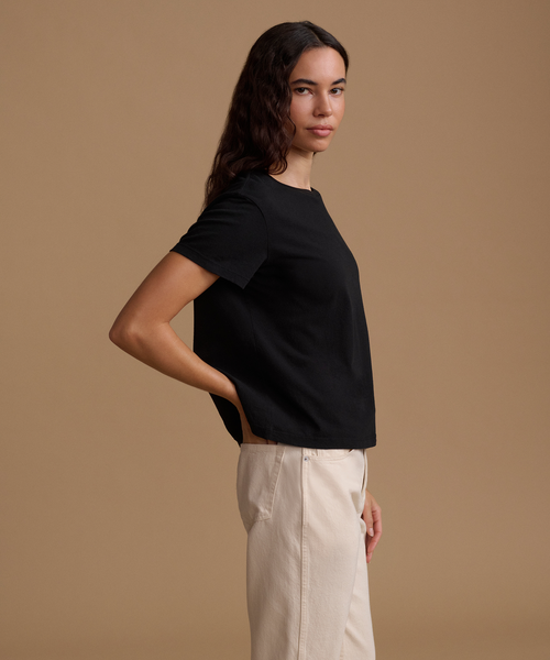 A woman modeling a black short-sleeve crewneck t-shirt paired with off-white trousers, standing against a solid warm beige studio background.