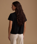 A woman with long, wavy dark brown hair is shown from the back, wearing a black short-sleeve t-shirt and cream-colored pants, set against a solid warm beige studio background.