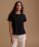 A woman modeling a black short-sleeve crewneck t-shirt paired with light beige pants, set against a solid warm brown studio background.