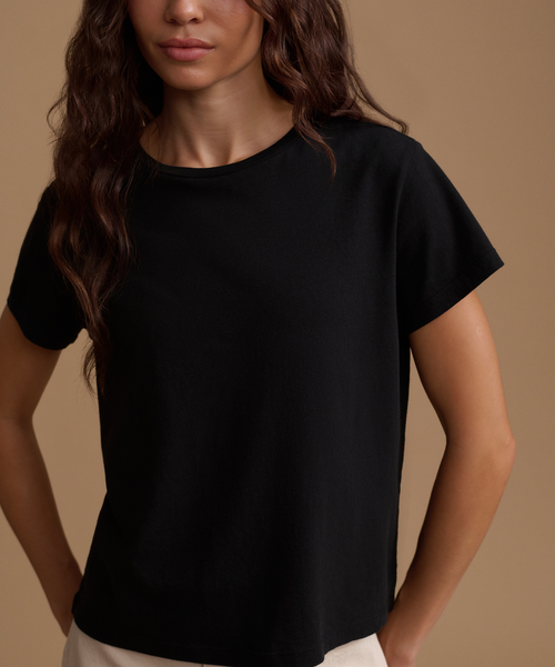 A woman wearing a plain black short-sleeve crewneck t-shirt, shown from the chest down against a solid warm beige background.