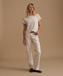 A model wearing a cream t-shirt with cream jeans and black leather shoes against a beige studio background.