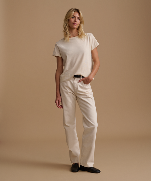 A model wearing a cream t-shirt with cream jeans and black leather shoes against a beige studio background.