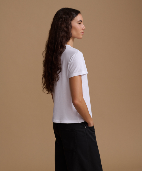 A woman with long, dark wavy hair is shown from a side-back angle, wearing a white short-sleeve crewneck t-shirt and black wide-leg pants, set against a solid warm tan studio background.