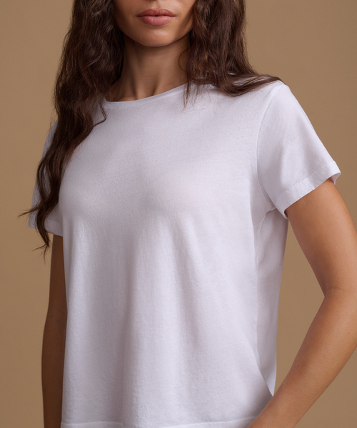A woman wearing a plain white short-sleeve crewneck t-shirt, shown from the chest up against a solid warm beige background.