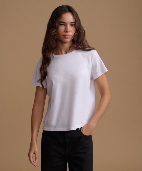 A woman modeling a white short-sleeve crewneck t-shirt against a solid tan studio background.