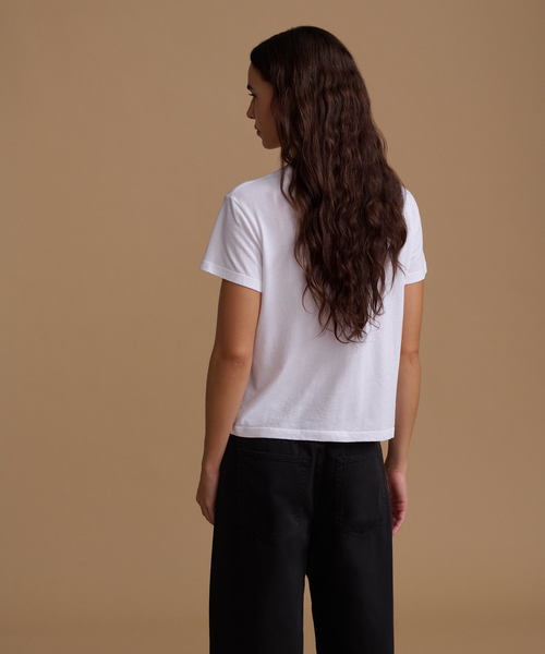 A woman with long, wavy dark brown hair is shown from behind wearing a plain white short-sleeve t-shirt and black wide-leg pants, standing against a solid warm beige studio background.