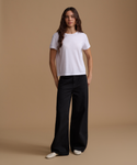 A woman stands against a plain warm beige studio background wearing a plain white short-sleeve crew-neck T-shirt, black wide-leg pants, and white pointed shoes.